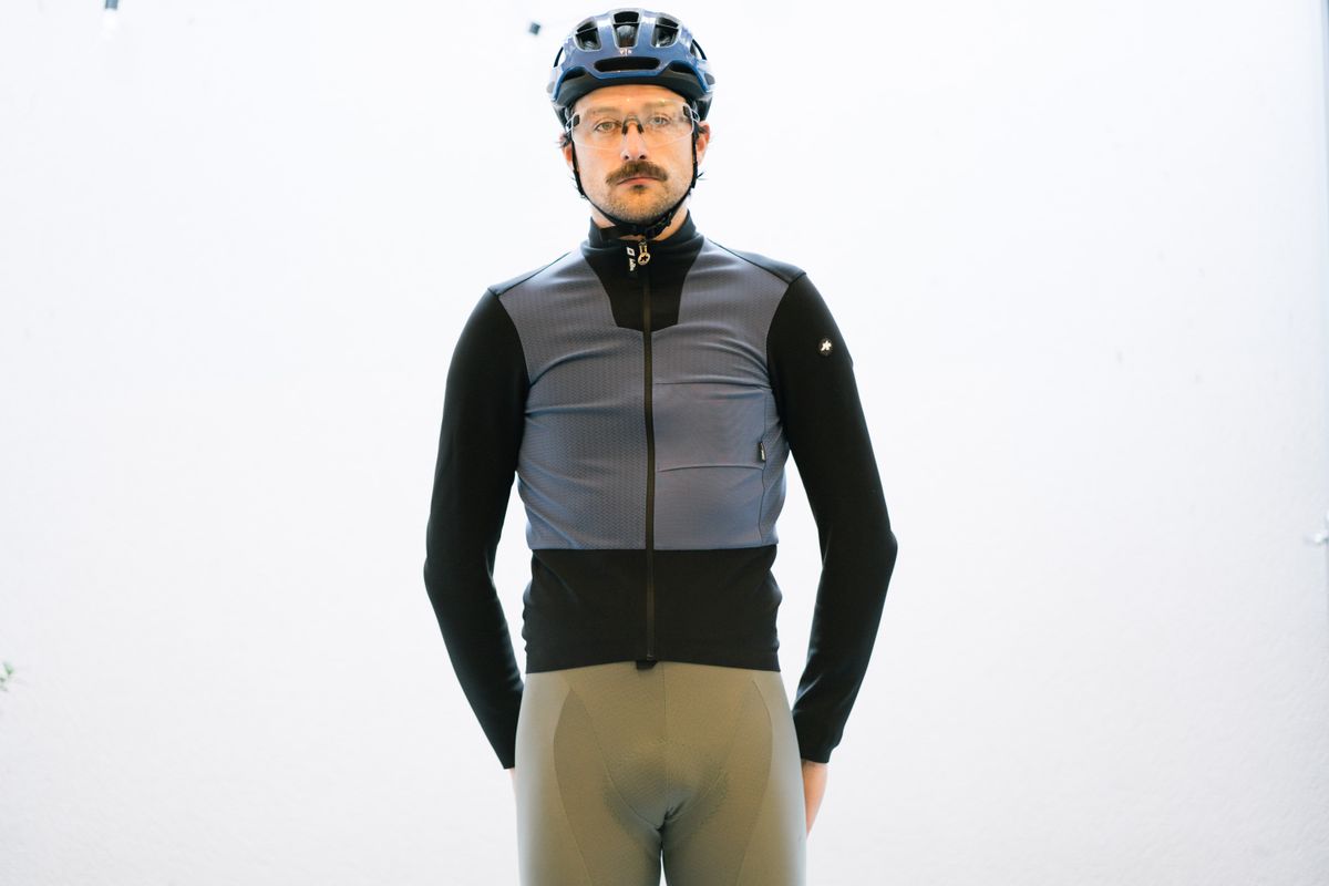 A white man standing in front of a white wall wearing a blue and black winter cycling jacket