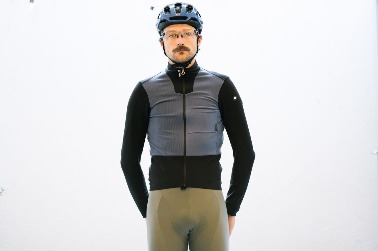 A white man standing in front of a white wall wearing a blue and black winter cycling jacket