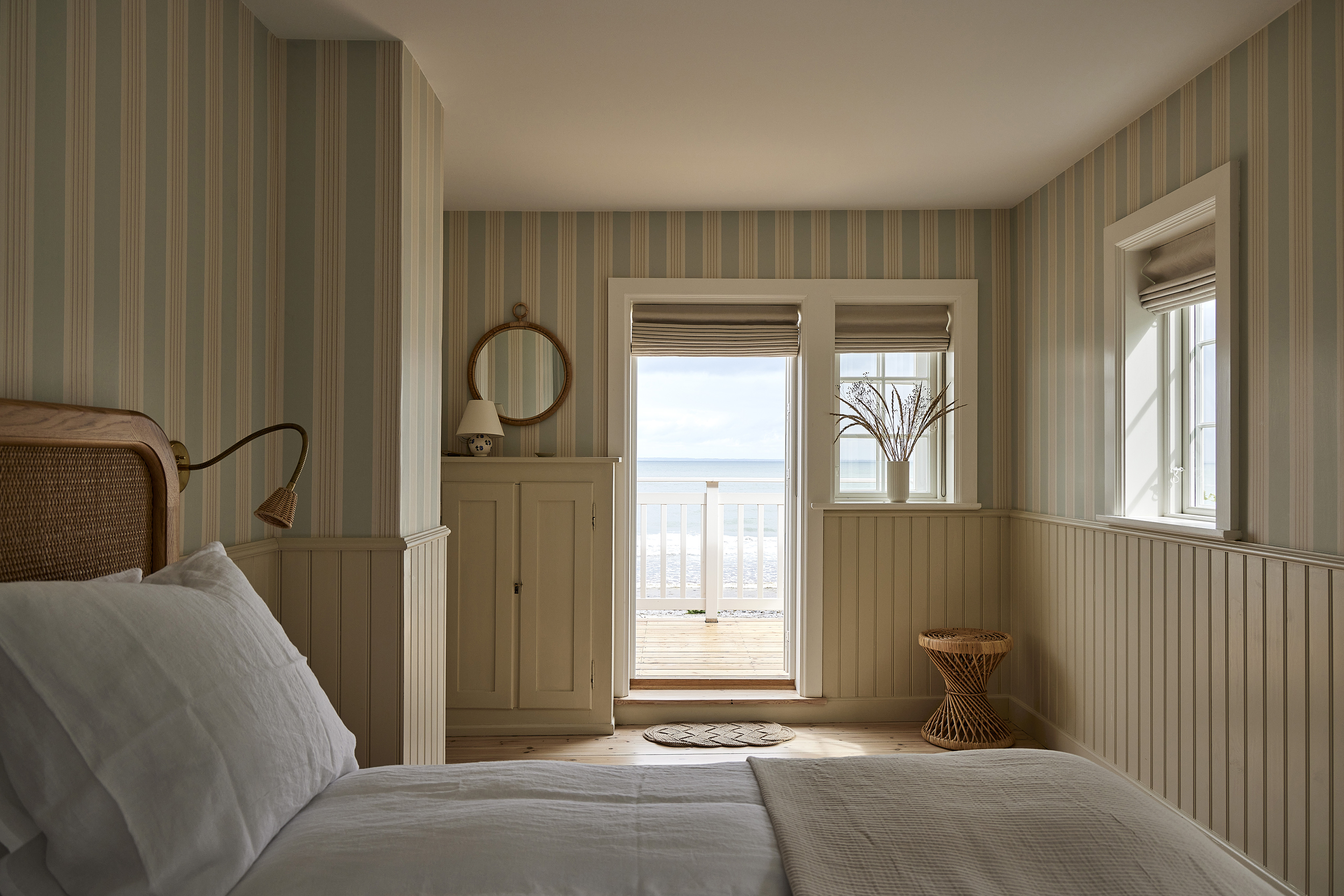 scandinavian coastal bedroom with striped wallpaper, wall paneling, white bed, rattan headboard, rattan stool, rope mirror above a cabinet with lamp and view to beach outside