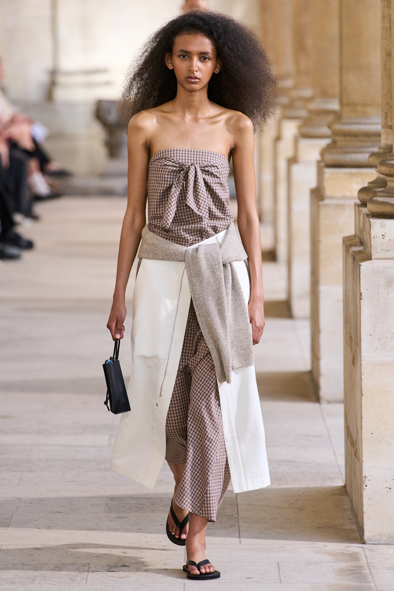 Layering staples for spring 2026 are shown in a photo from Auralee's runway show, with a model walking while wearing a brown and white gingham tube top styled with matching trousers, a white skirt, and a tan sweater layered on top, paired with black flip-flops and a black mini bag
