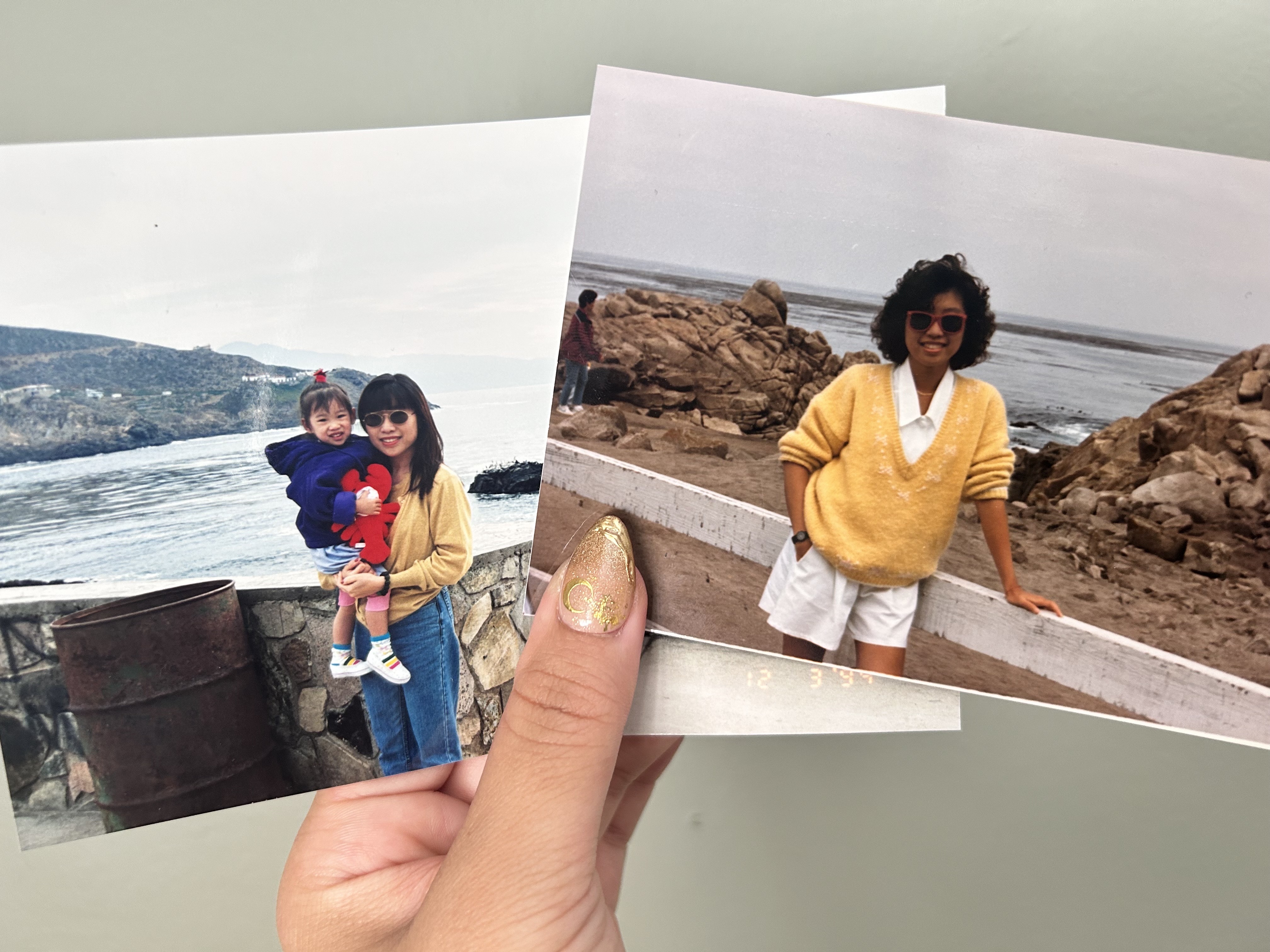 A hand holding two print photos. On the left, a toddler is being held by her mother; on the right, the mother is posing in a yellow sweater and white collared button-down with white shorts.