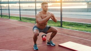 Man performing a squat outdoors next to exercise mat and kettlebell