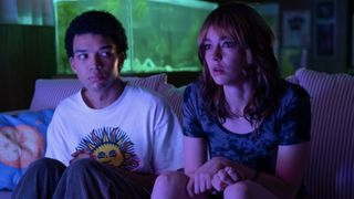 Two teenagers sit on a couch with a fish tank in the background