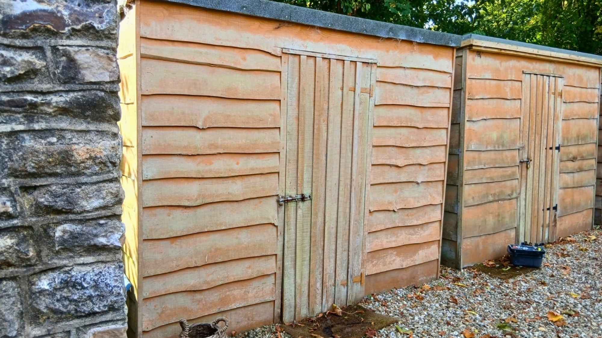 Two wooden garden sheds with latched doors beside a stone wall, on a gravel surface.