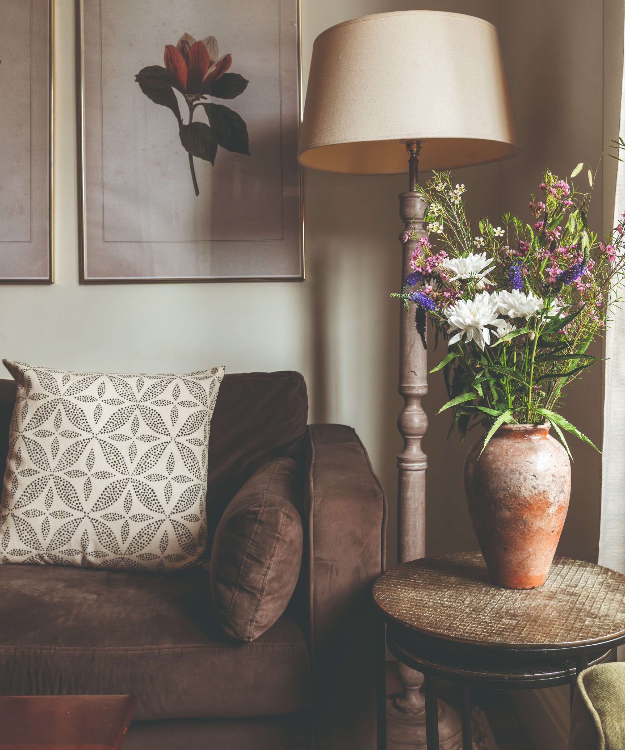 A living room with a brown sofa and a rustic floor lamp and vintage side table next to it with a vase of flowers on top