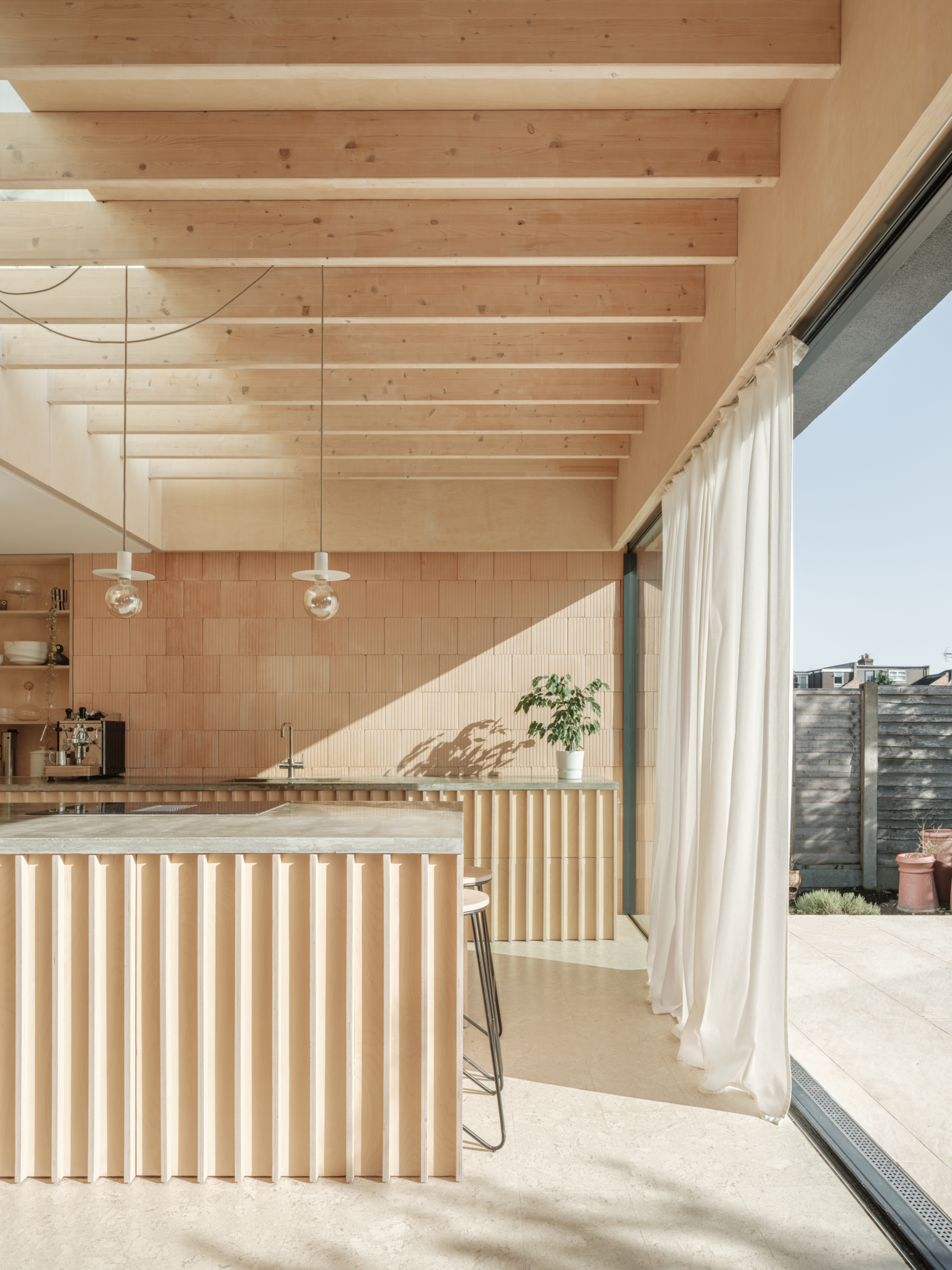 Open-plan kitchen extension with exposed beams roof, exposed clay block wall, and clay fluted island with white sheer curtains half covering the large door frames leading to the outside space