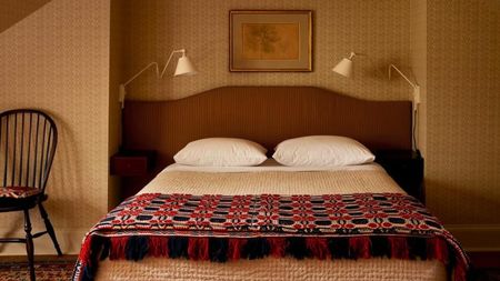 Image of a brown bed with cream bedding and a red and black throw in a room that is covered in caramel wallpaper.
