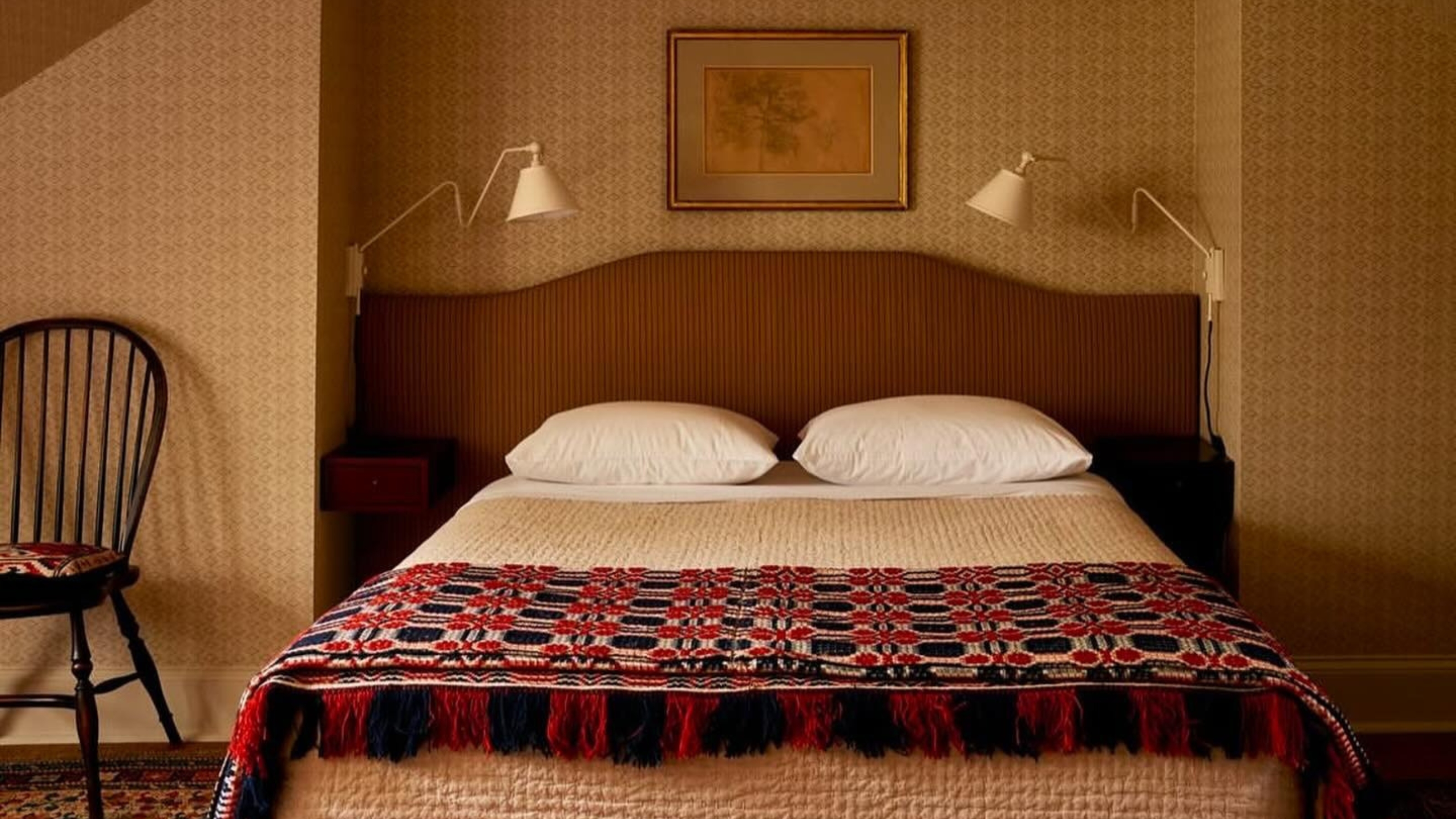 Image of a brown bed with cream bedding and a red and black throw in a room that is covered in caramel wallpaper.