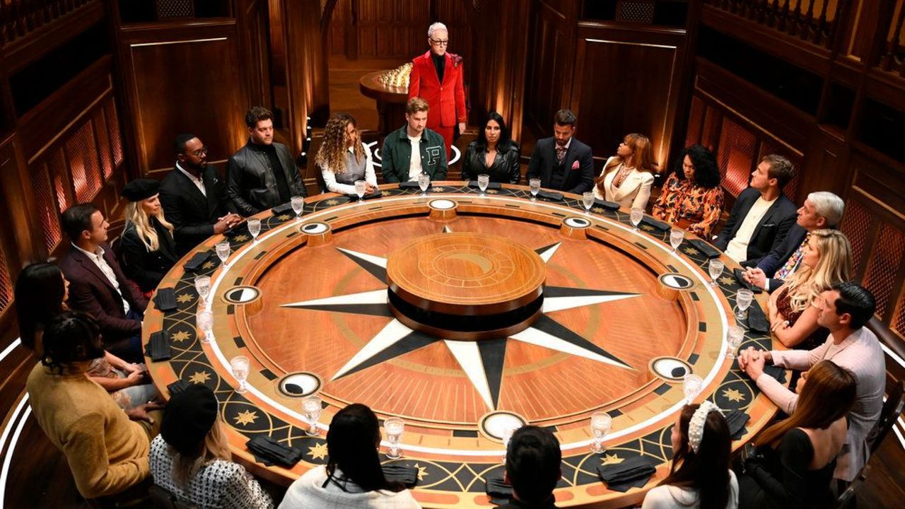 a group of people sit around a round table decorated with a star motif, as alan cumming stands among them wearing a red suit, in &#039;the traitors&#039;