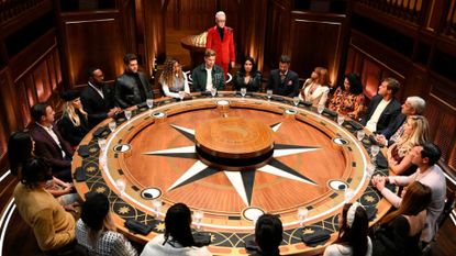 a group of people sit around a round table decorated with a star motif, as alan cumming stands among them wearing a red suit, in 'the traitors'