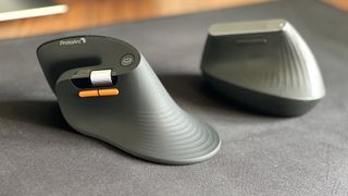 Side-by-side view of the ProtoArc EM11 Pro ergonomic mouse in the foreground with its orange side buttons and scroll wheel, next to the Logitech MX Vertical mouse sitting on a black desk mat.