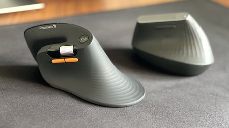 Side-by-side view of the ProtoArc EM11 Pro ergonomic mouse in the foreground with its orange side buttons and scroll wheel, next to the Logitech MX Vertical mouse sitting on a black desk mat.
