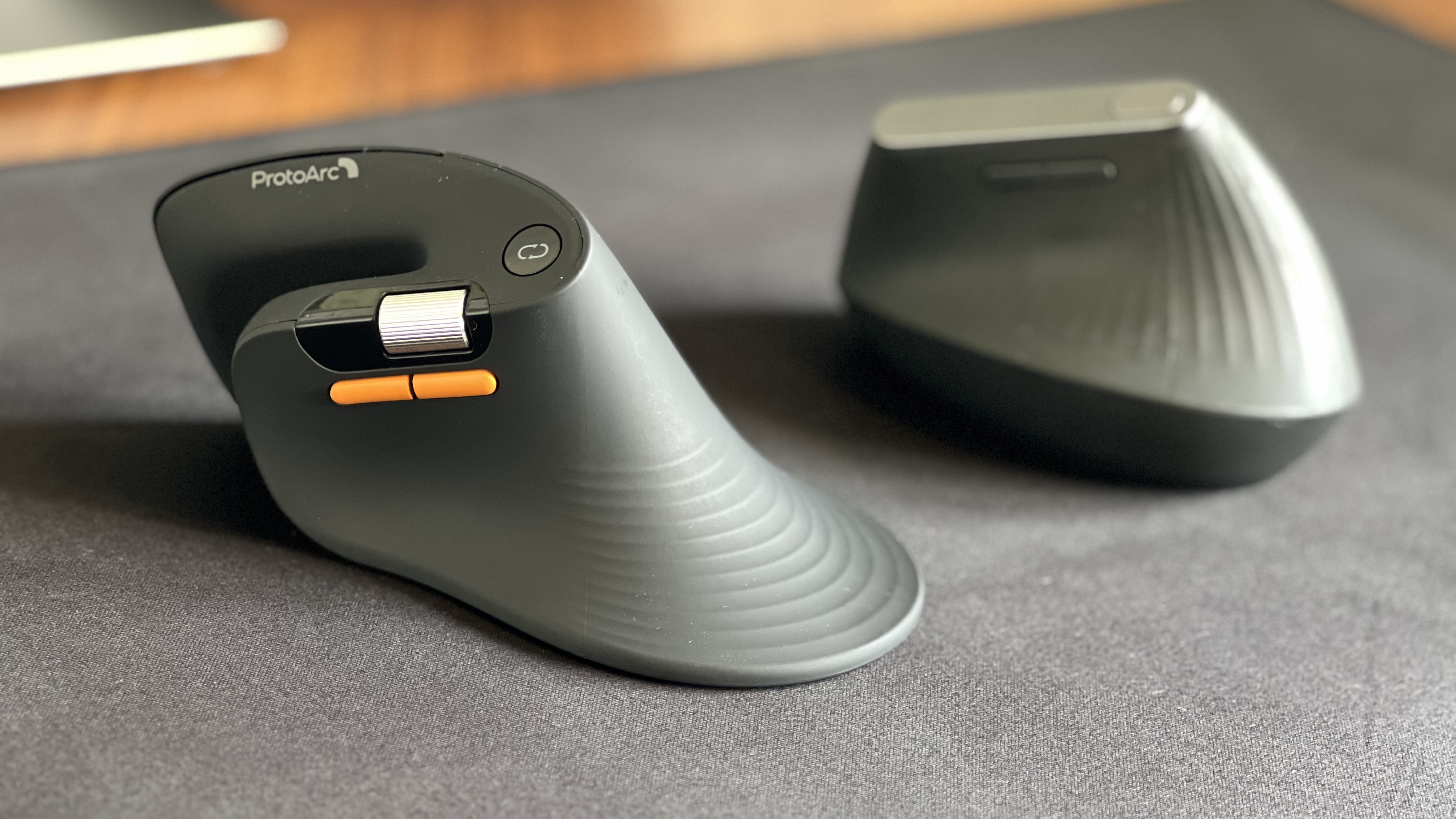 Side-by-side view of the ProtoArc EM11 Pro ergonomic mouse in the foreground with its orange side buttons and scroll wheel, next to the Logitech MX Vertical mouse sitting on a black desk mat.