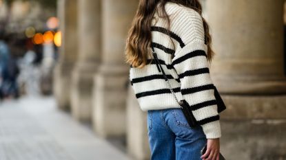 A passerby wears a white with black striped oversized pullover, a gold ring, blue denim large jeans pants, a black shiny leather crossbody bag, on November 05, 2021 in Paris, France. (Photo by Edward Berthelot/Getty Images)