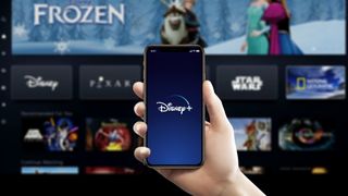 Disney Plus logo on a right-hand held smartphone with the app loaded on a TV in the background