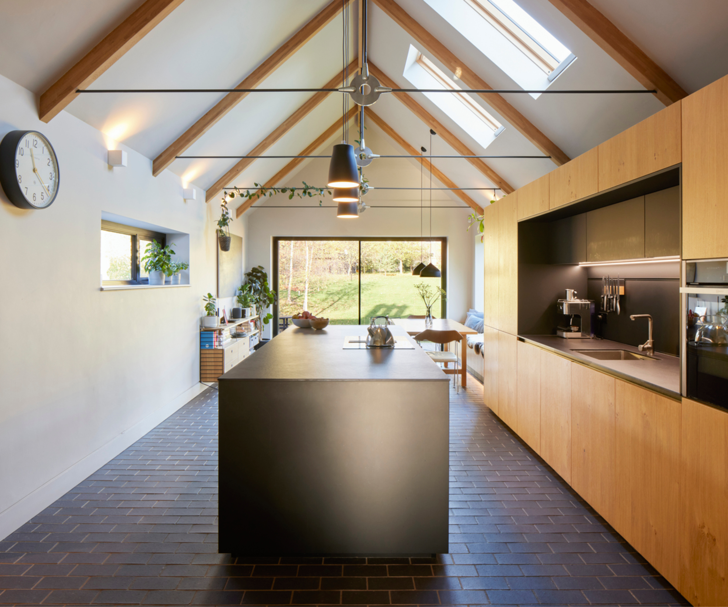 single storey kitchen extension with large vaulted ceiling showing modern glulam beams, wooden floor to ceiling handleless kitchen units and dark central island with pendant lighting above