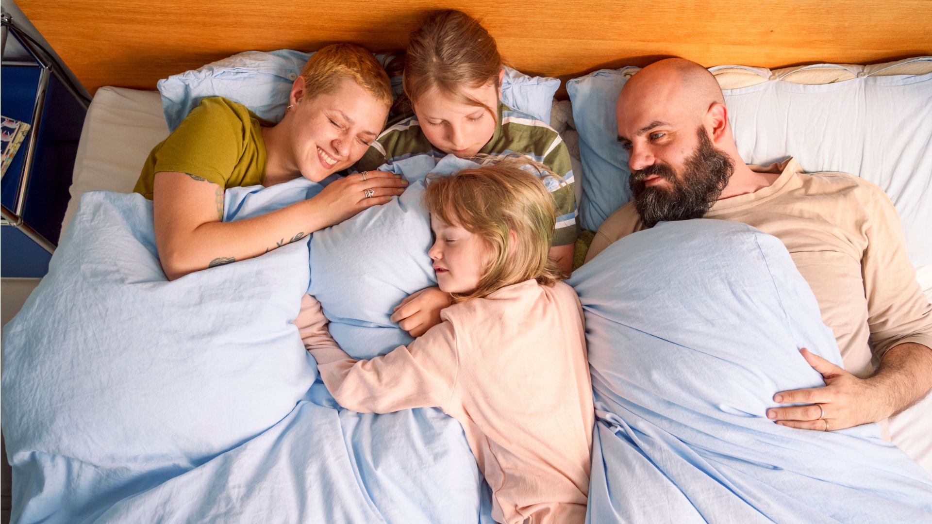 A family of four cuddled up in bed together. The parents are awake but both the children are sleeping.