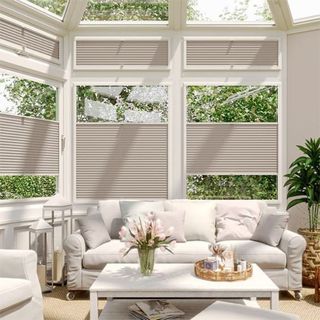 A bright conservatory with white-framed windows and a glass ceiling. Beige pleated blinds cover the upper and lower sections of the windows, letting in soft natural light. A white sofa with plush cushions sits against the windows, accompanied by a matching armchair. 