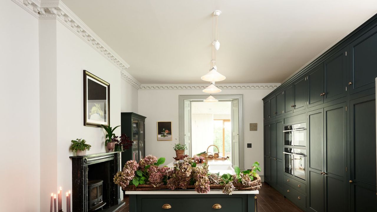 Converted Victorian home&#039;s large galley kitchen. Dark green cabinets on the right with double oven tower stack, island in middle, fireplace on left. Brass handles and hardware with crown molding on ceiling