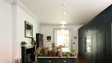 Converted Victorian home's large galley kitchen. Dark green cabinets on the right with double oven tower stack, island in middle, fireplace on left. Brass handles and hardware with crown molding on ceiling