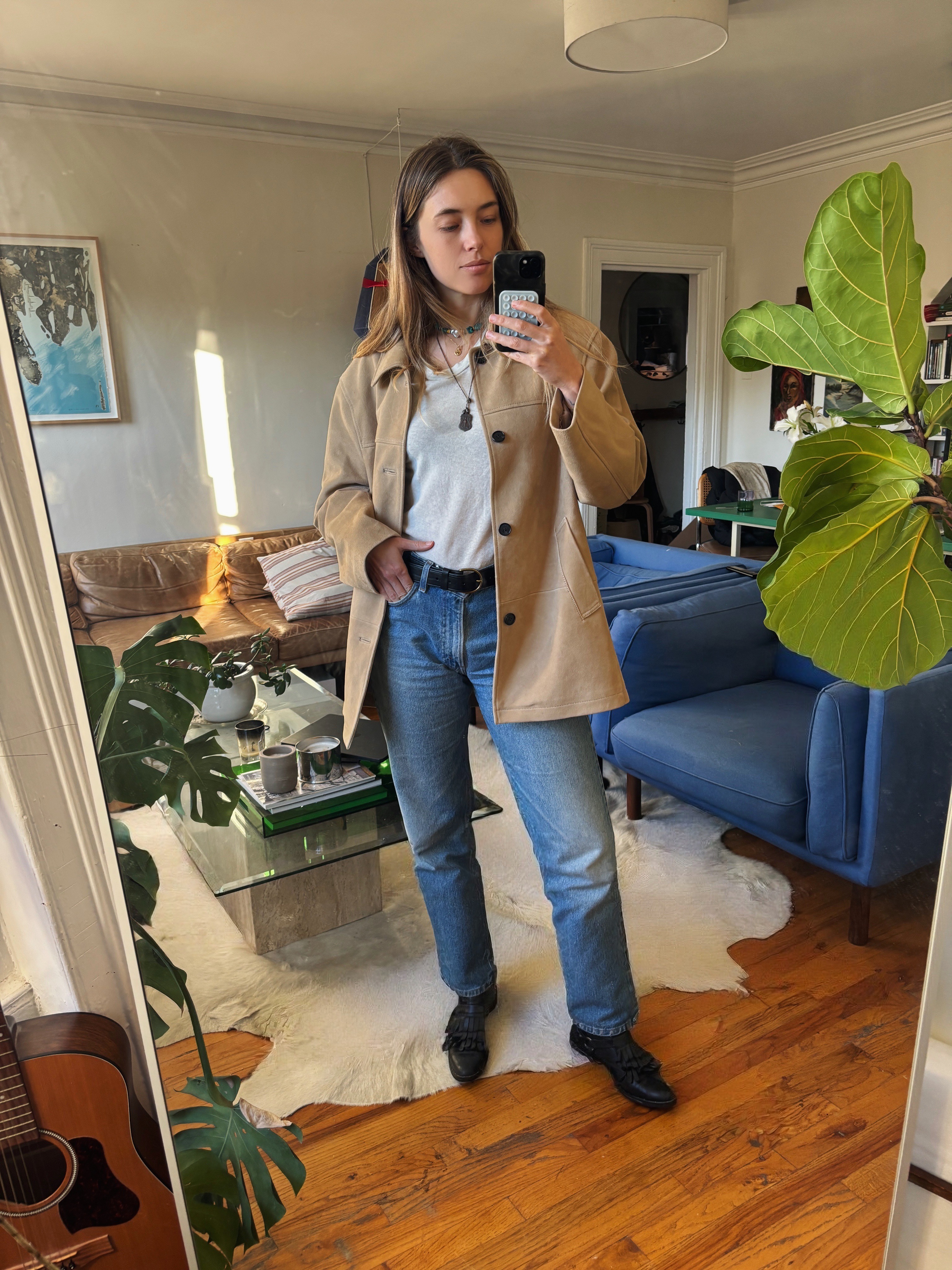 Aemilia Madden taking a selfie, wearing a tan work coat, blue denim jeans, a brown belt, brown shoes, and silver necklace, holding an iPhone