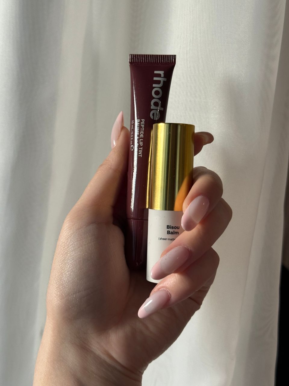 An image showing Denise Primbet holding the Violette FR Bisou Balm and the Rhode Peptide Lip Tint