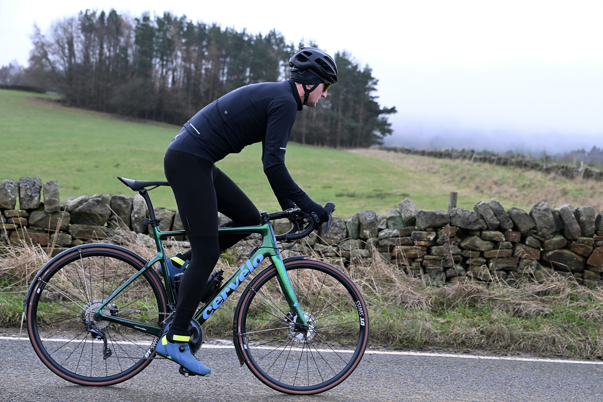 side view of a man riding a green Cervelo road bike wearing black tights, black jacket, black helmet and blue shoes