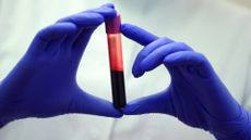 A person wearing all white holds a plastic tube with a pink cap half full of reddish liquid in their two hands covered in purple nitrile gloves. 