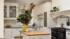 A small kitchen with glass cabinets, open wall-shelves, a gas stove, a moka pot, a wooden island with a vase of stems