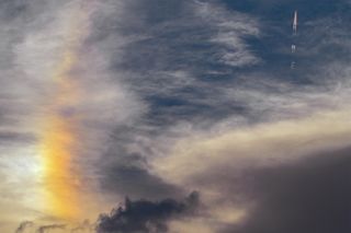 Partly cloudy sky with a rainbow sundog on the left and a plane with rainbow contrail in the upper right corner of the image.