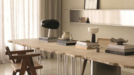 close up of stone dining table with textured edge, cream wall with stainless steel shelving, and armchair. the table is stacked with coffee table books with lamp