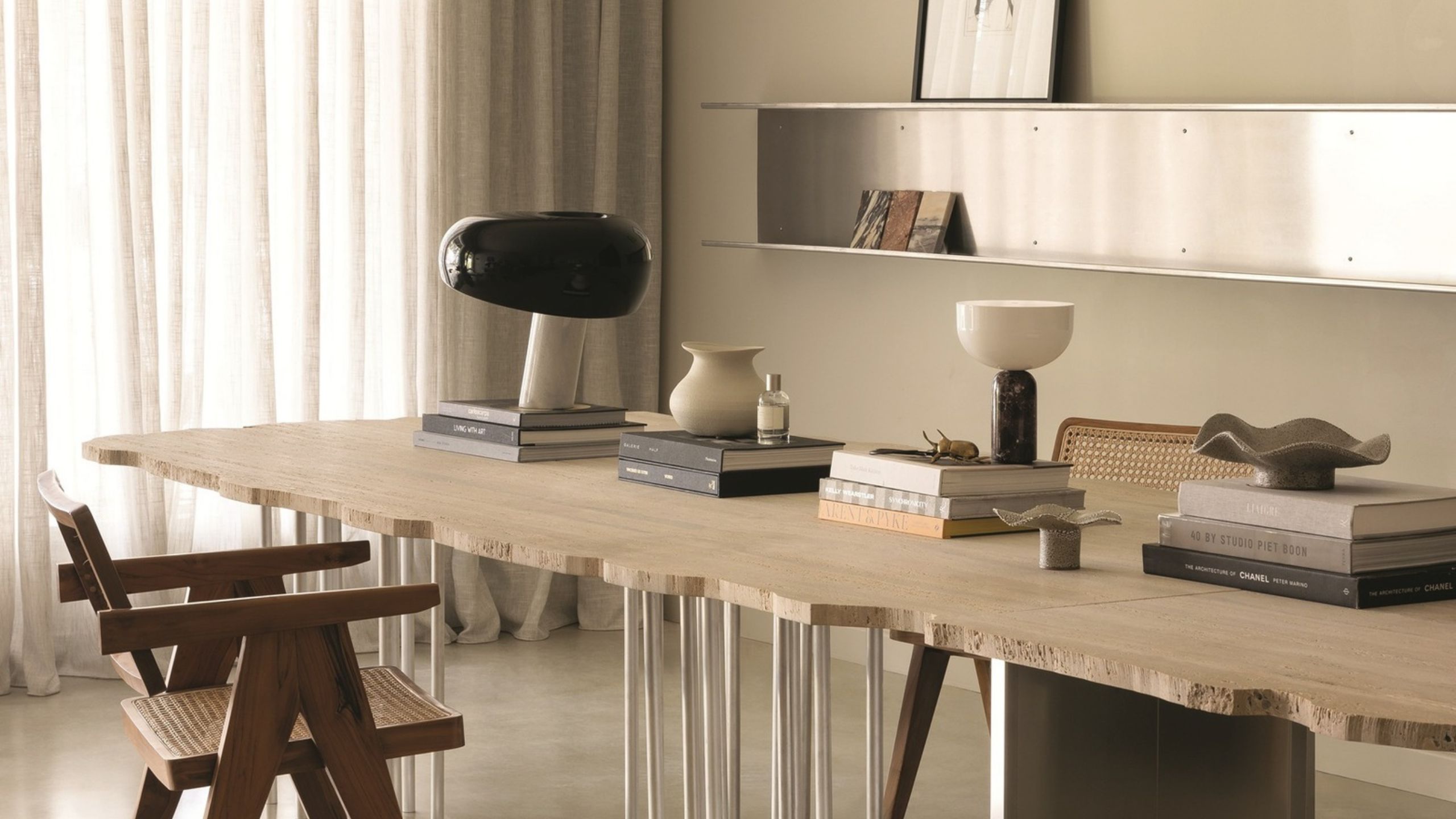 close up of stone dining table with textured edge, cream wall with stainless steel shelving, and armchair. the table is stacked with coffee table books with lamp