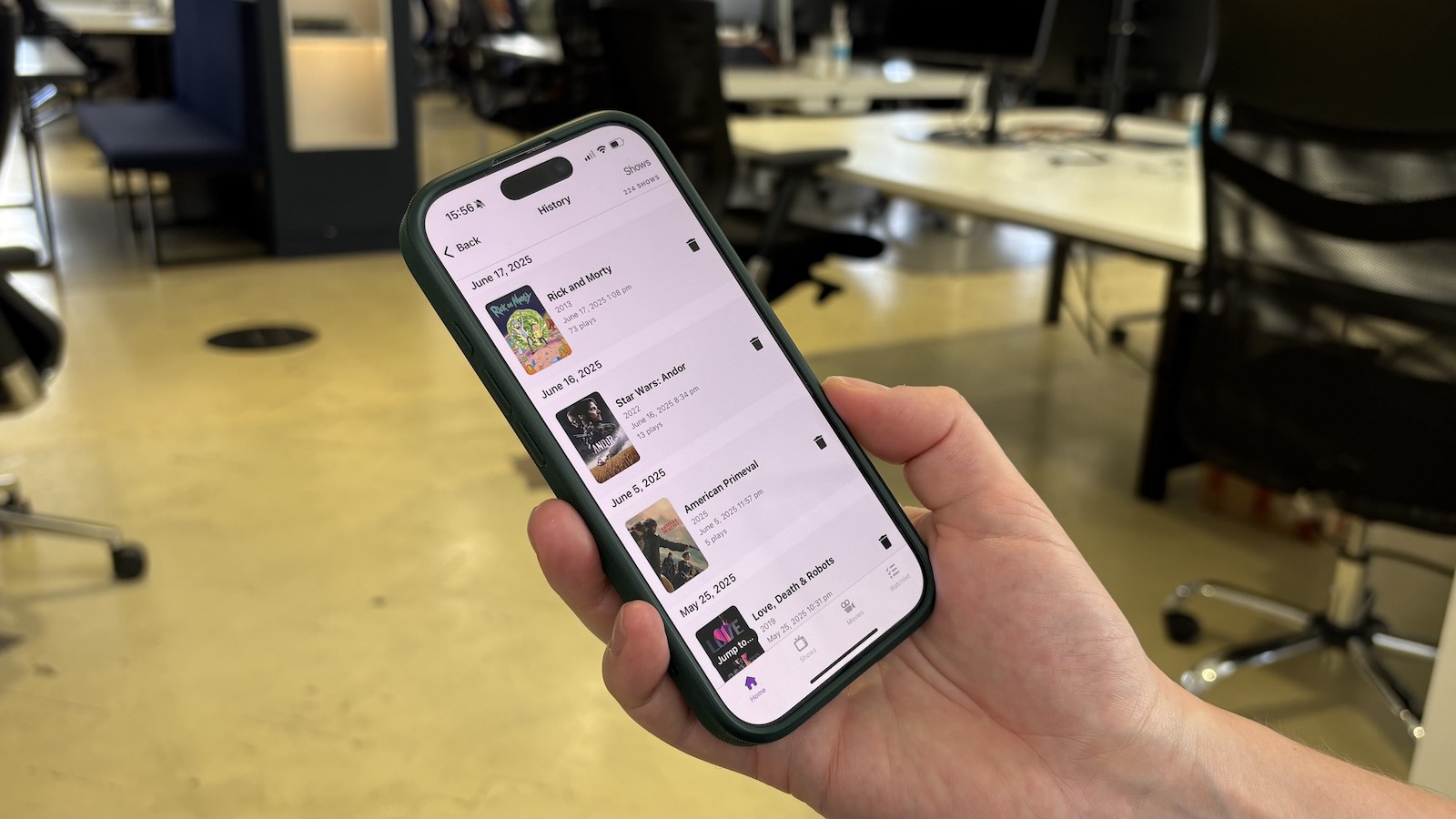A photograph of someone holding an iPhone. On the screen is the history section of the Trakt app.
