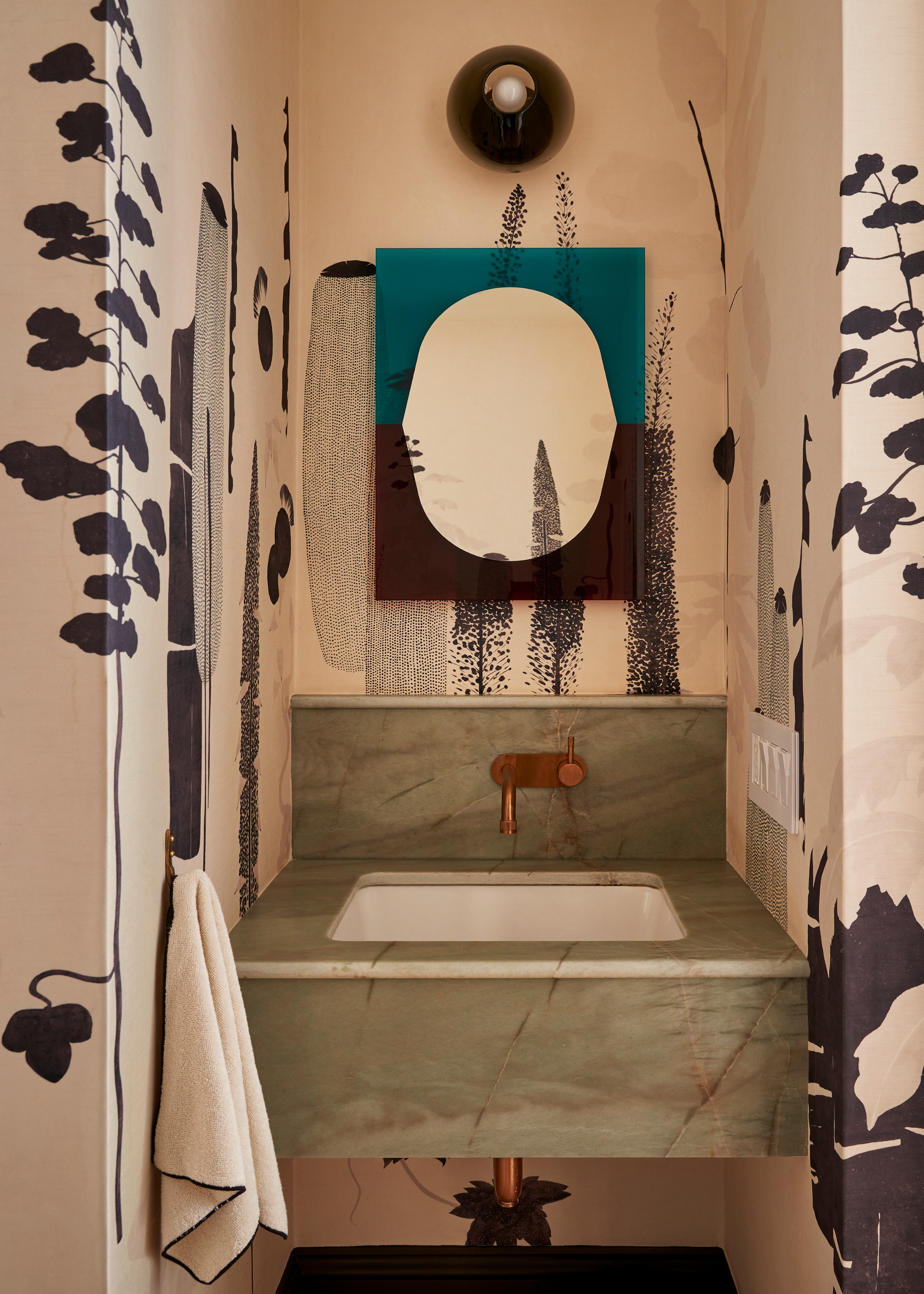 A powder room with a green marble basin, bronze hardware, wallpaper, a black sconce, and a colored mirror