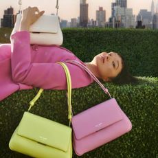 Model wearing a pink suit jacket with 3 Kate Spade bags lying on a hedge