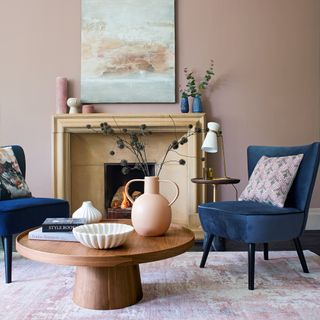 pink living room with blue velvet armchiars and round wooden coffee table in front of a fireplace