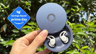 The image shows a woman's hand holding a pair of white earbuds in their navy blue case in front of a green plant. Inset to the left, a deal badge reads 'Tom's Guide Saving Squad Prime Day'