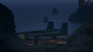 Still of the Exile ship from the high republic era of Star Wars in a landing position on a beach with the sea in the background and some lights from buildings to the side.