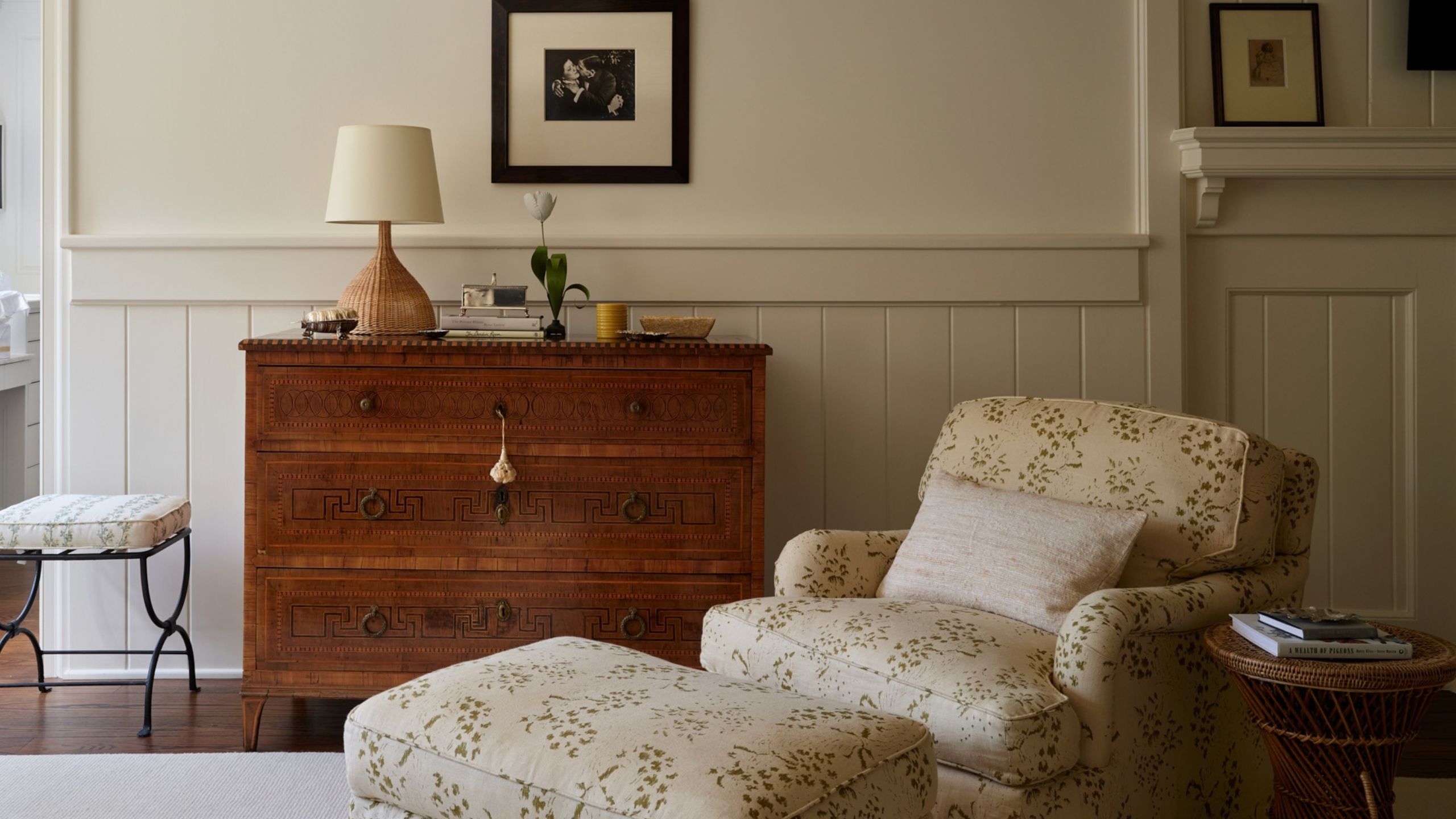 a cozy warm neutral bedroom with half-height wainscoating styled with an antique dresser, green and yellow floral armchair and matching ottoman and small side tables and stools