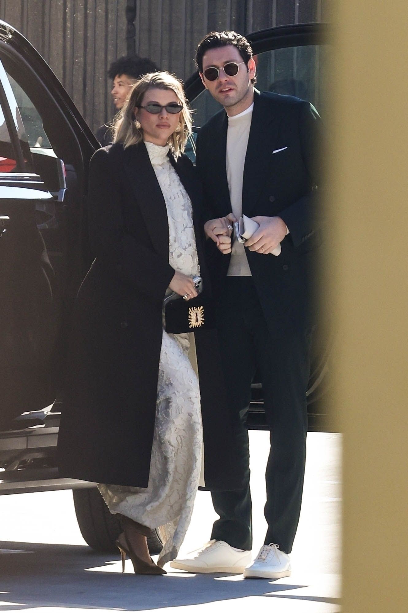 Sofia Richie wears a Schiaparelli two piece skirt set with a black coat, black bag and gold shoes.