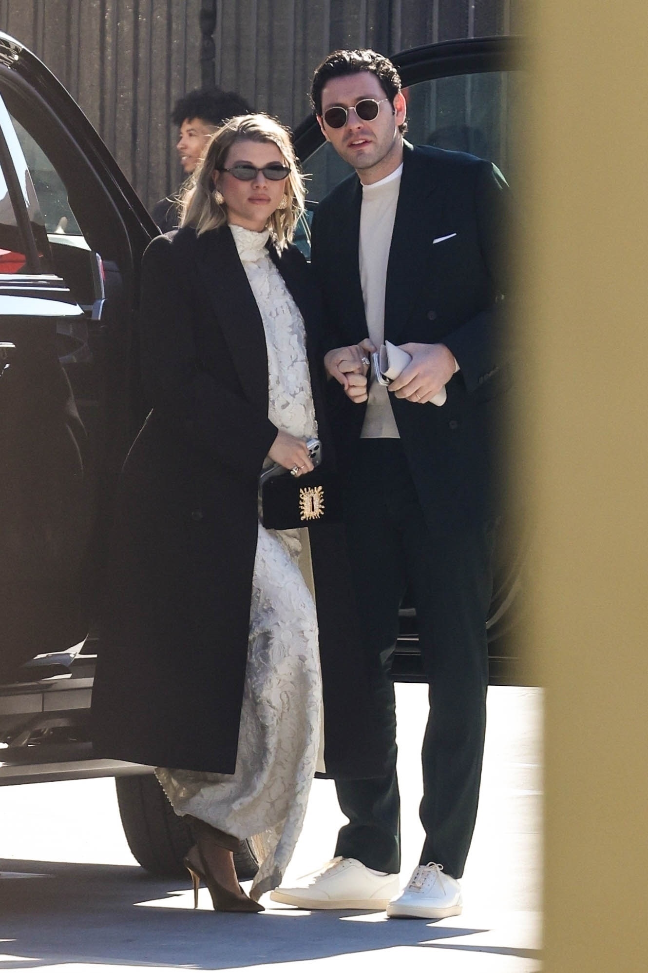 Sofia Richie wears a Schiaparelli two piece skirt set with a black coat, black bag and gold shoes.