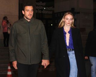 Sophie Turner wearing an open blue button down with a black wool coat and light wash jeans