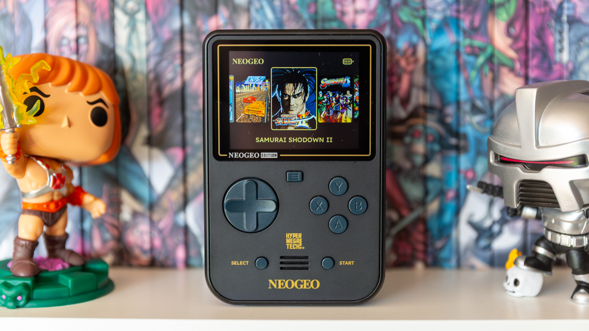 HyperMegaTech! Neo Geo Super Pocket on a shelf in front of 2000AD books and flanked by two Funko Pops
