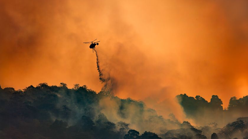 A photograph of a helicopter dropping water on a forest fire in Thailand. 