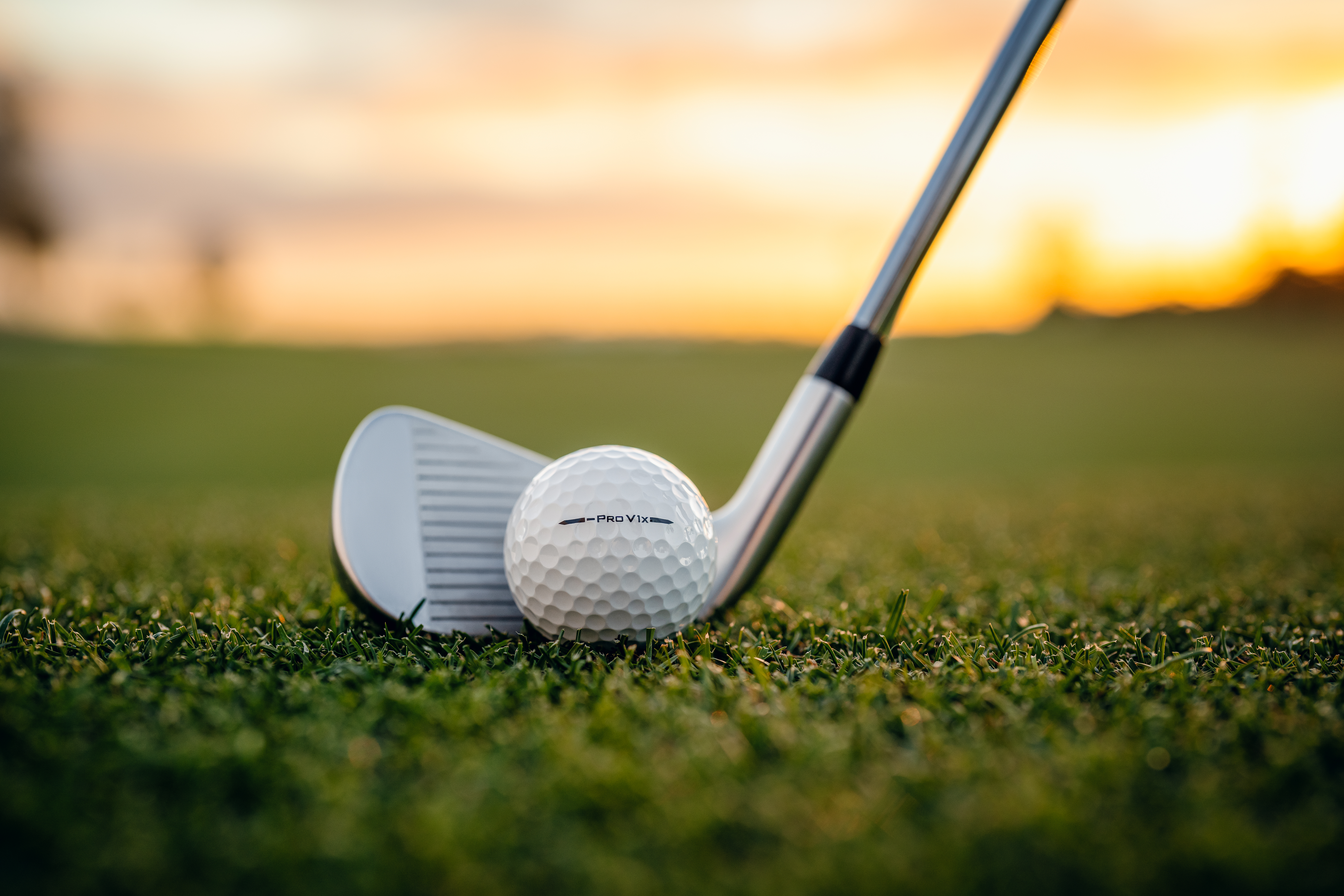 Close-up of a golf club addressing a dimpled golf ball on short grass, with a soft-focus fairway and warm sunset sky in the background.