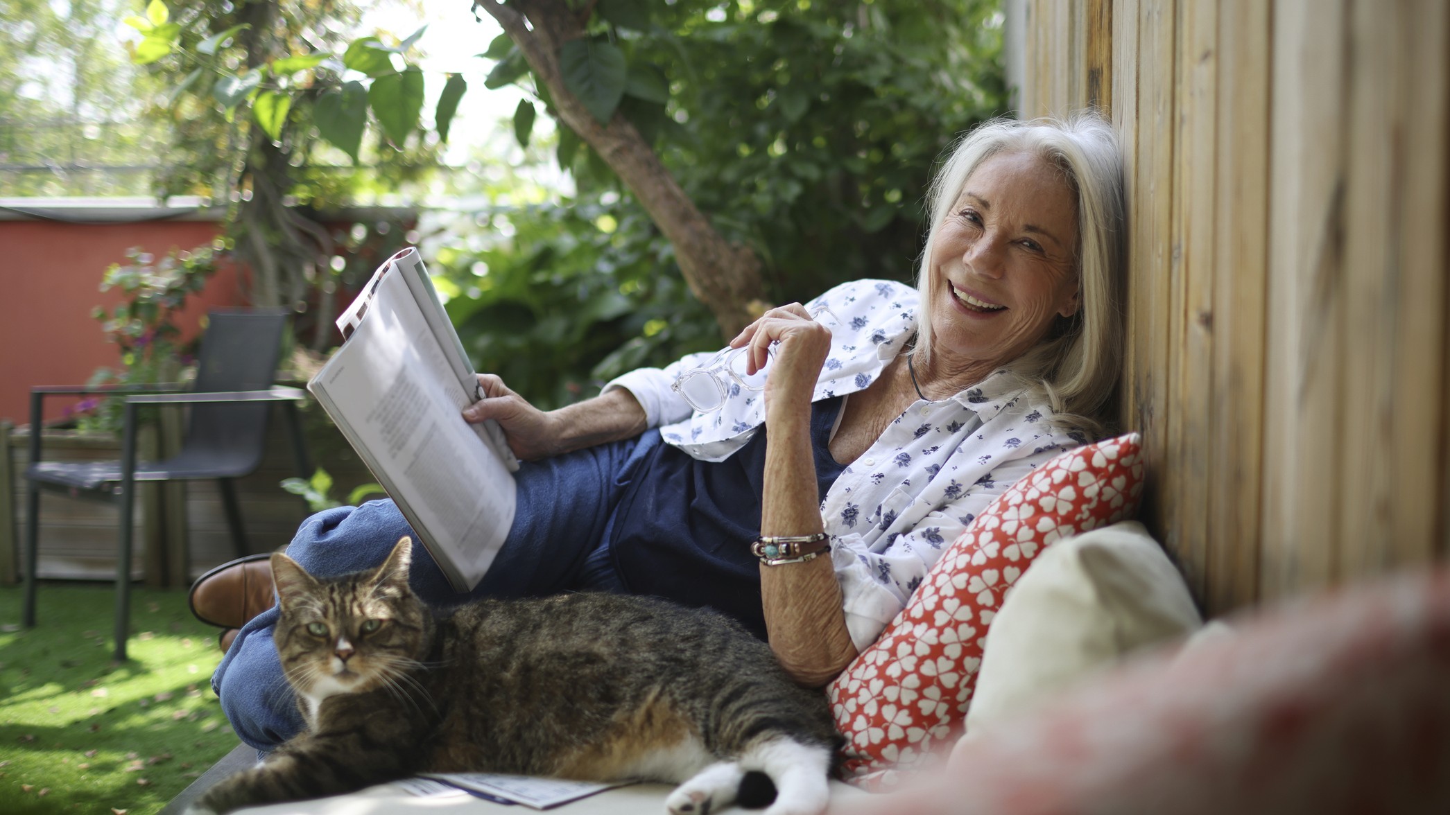 A woman with grey hair reads a packet outside with a cat, smiling. 