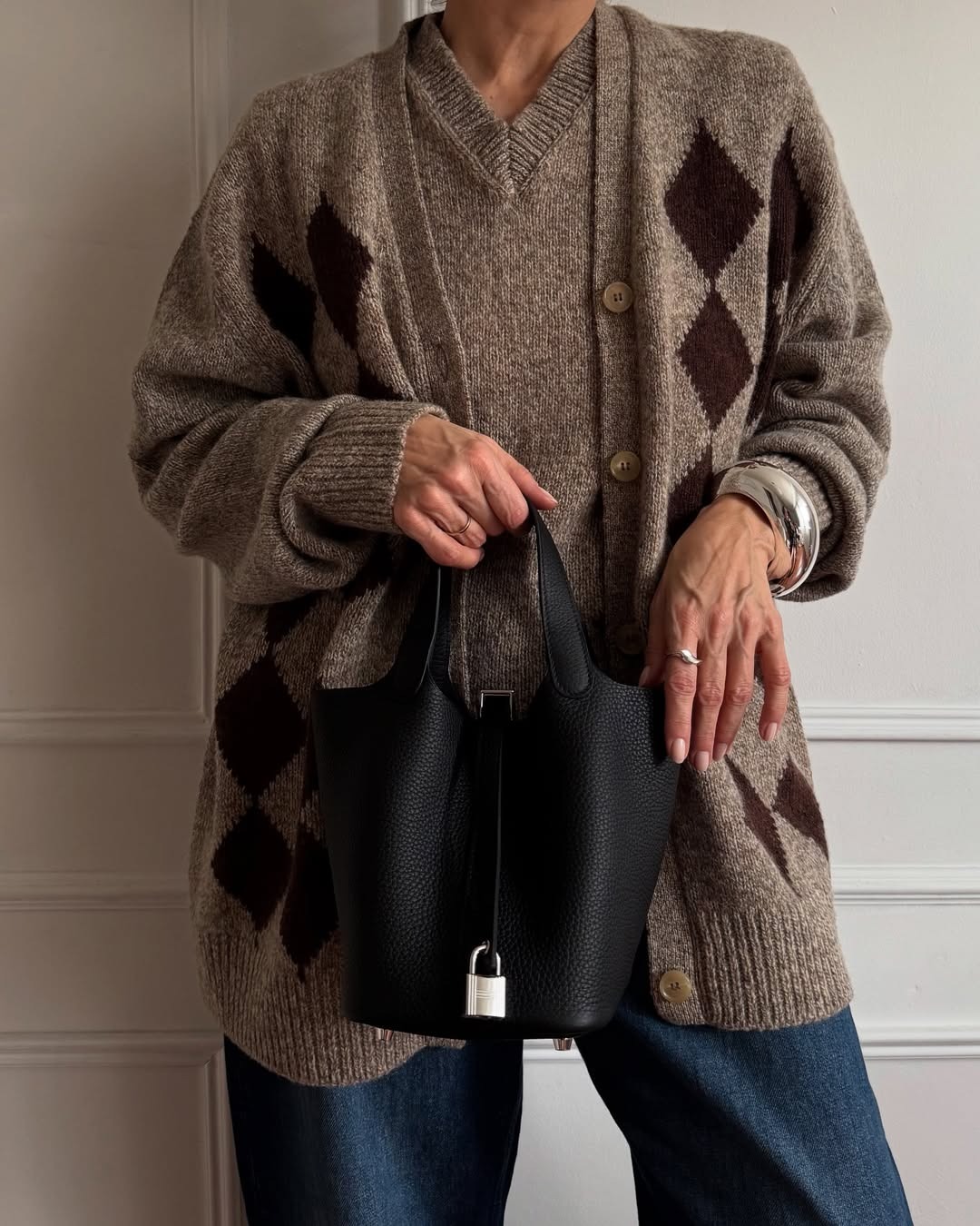 Renia Jaz holding a black Hermes handbag with lock detailing against a brown argyle sweater.