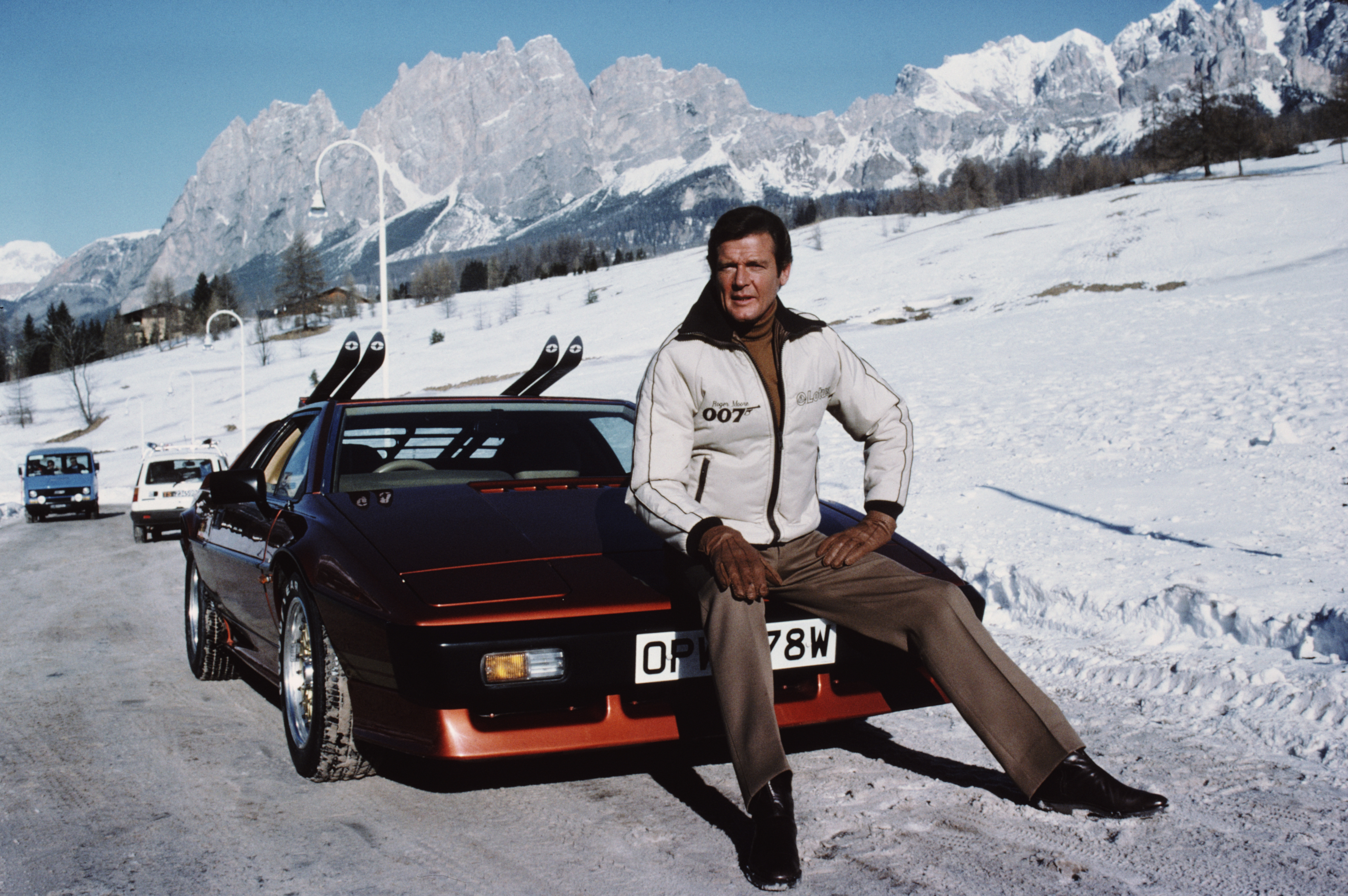 Roger Moore as James Bond, sitting on top on a car with skis on top in the snowy mountains