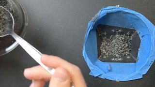 a person moves gray gravel from a blue cup to a glass one using a spoon