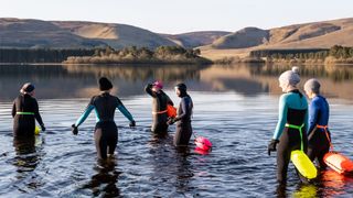 A group of women friends gathering together at the start of the day to wild swim in a Scottish freshwater reservoir, with hills surrounding the water. They are wearing wetsuits and warm hats to protect themselves from the cold water. They are wearing brightly coloured inflatable swimming safety belts attached round their waists. The friends stay close together for safety and companionship as they exercise. It is a fun activity that helps with mental health, wellbeing and is enjoyable time out before returning to their families, work and the day ahead.